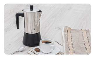 Cafetera italiana junto a una taza de café negro y un trozo de chocolate.